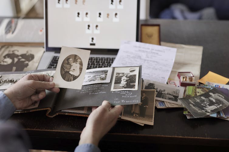 Someone whose face and body are not visible sits at a table, holding up black and white photos in front of an open laptop.