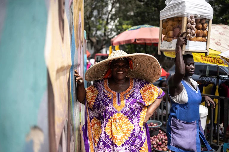 A smiling woman wearing a wide-brimmed hat. Another woman in the background with a container of goods on her head.