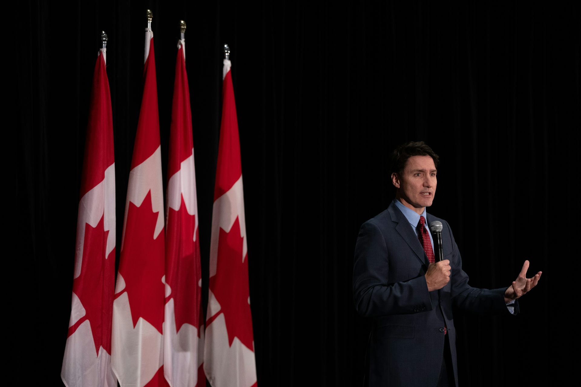 A dark-haired man gestures as he speaks into a micorphone with a row of Canadian flags behind him.