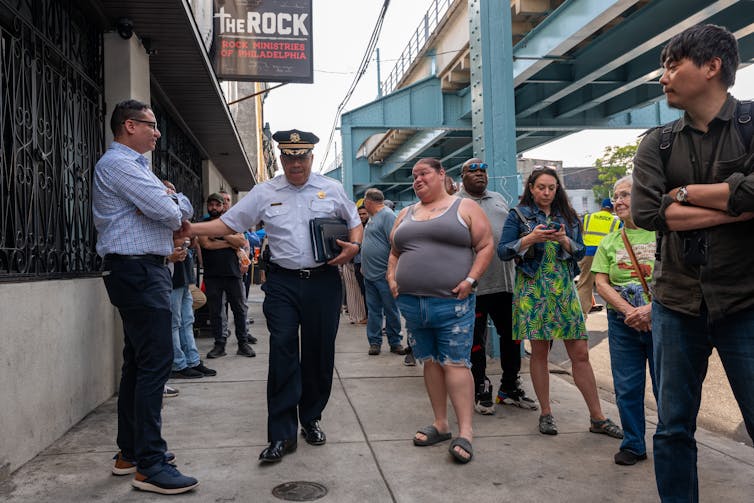 A uniformed police officer walks down a sidewalk with about a dozen people around him
