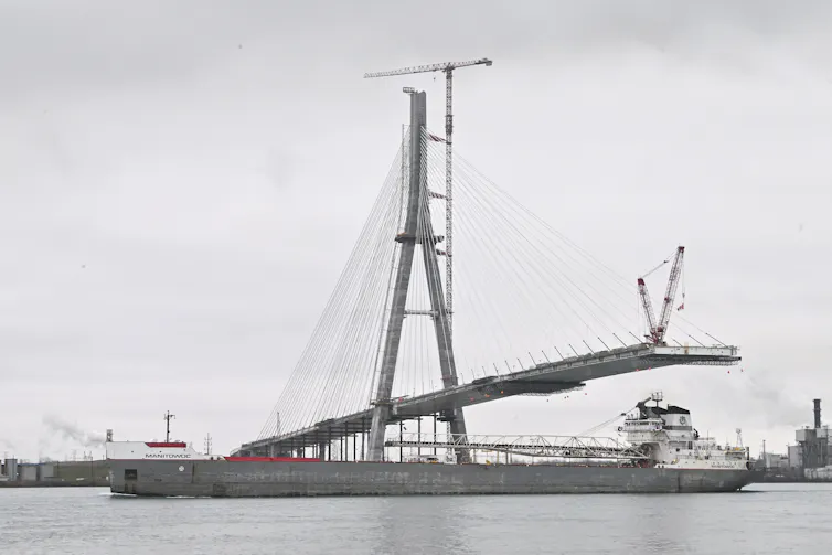 A large freighter sails past a suspension bridge under construction over a wide river.