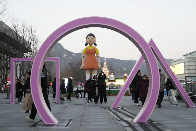 People walk past an art installation for Squid Games Season 2. It consists of a purple circle, square and triangle rising out of the ground in front of a giant doll with pigtails