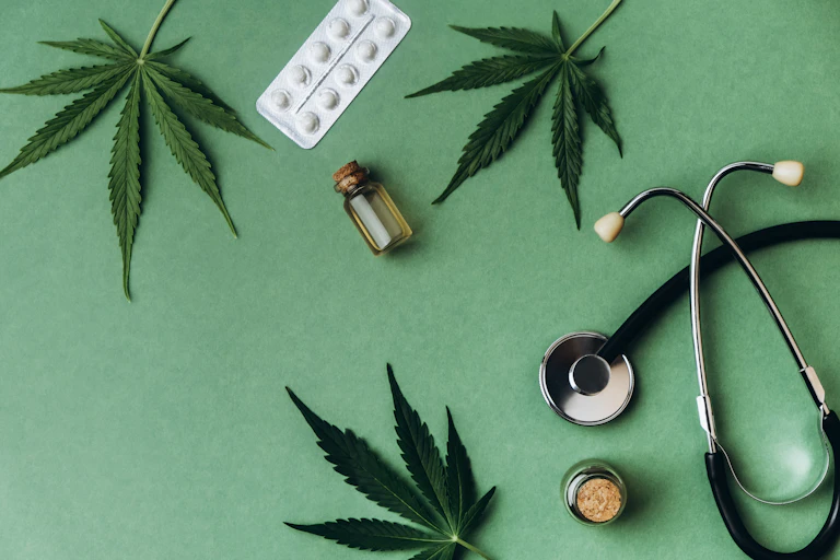 Three stems of cannabis leaves, a stethoscope, and a pack of white pills and tincture bottle lie on a green background.