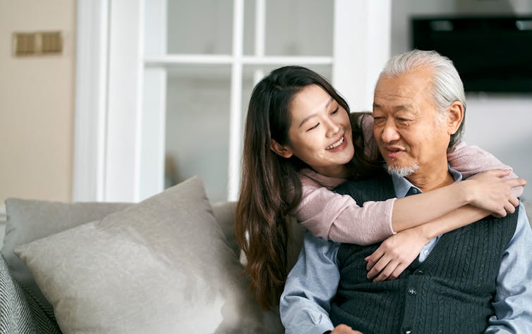 A young woman smiles with her arms around an older man seated on a sofa