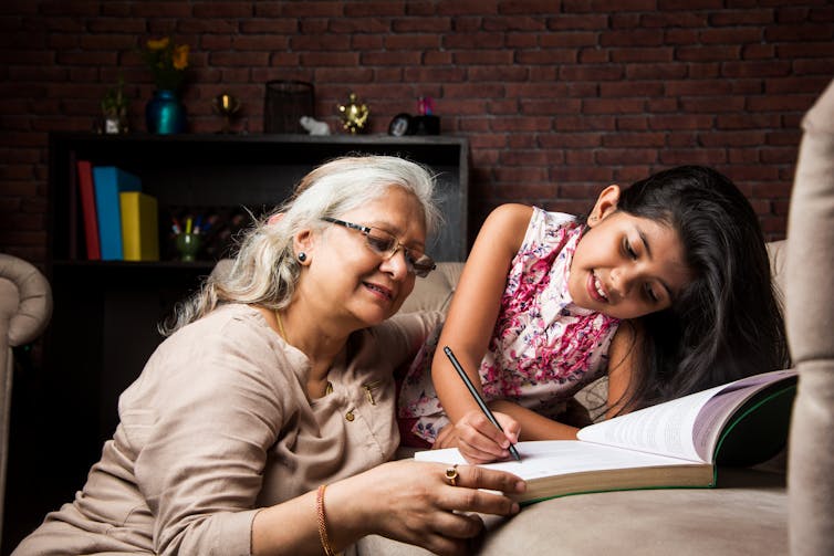 An older woman with grey hair helps a young girl seated on a couch do her homework.