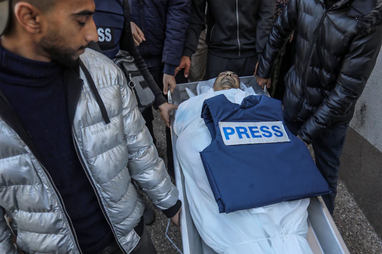 A deceased body, wrapped in a white shroud with the head exposed, is carried in an open coffin. A "press" vest is on top of his body, and he is surrounded by other people.