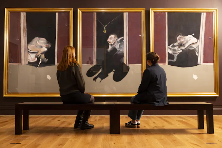 Two women sat in front of a triptych of Bacon paintings.