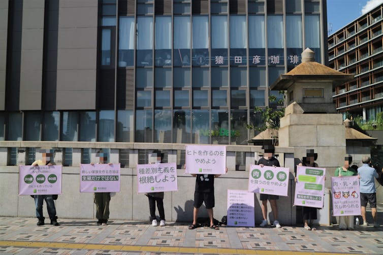 Seven people stand behind large placards they are holding outside on a city street.