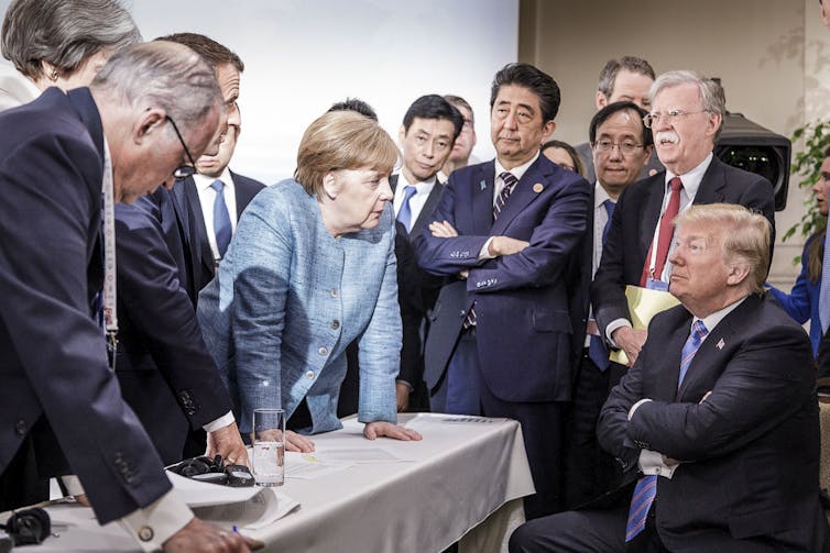 A woman wearing a light blue jacket stands around a table with men wearing dark suits. She faces toward a man seated with his arms crossed.