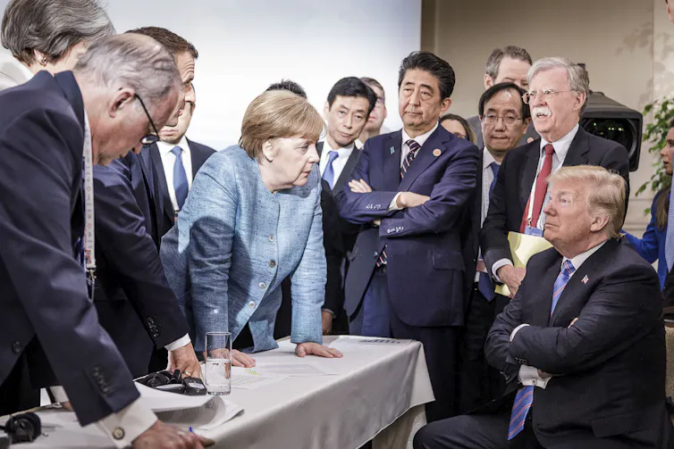 A woman wearing a light blue jacket stands around a table with men wearing dark suits. She faces toward a man seated with his arms crossed.