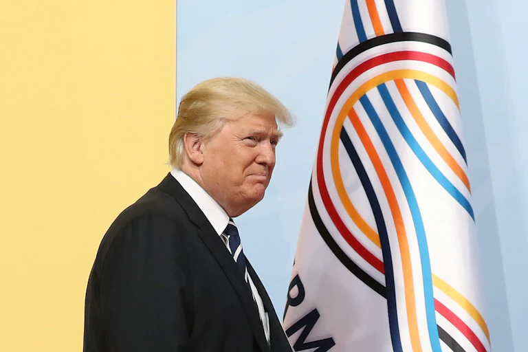An older man with yellow hair and a black suit walks past three flags, including one that is black, red and yellow striped.