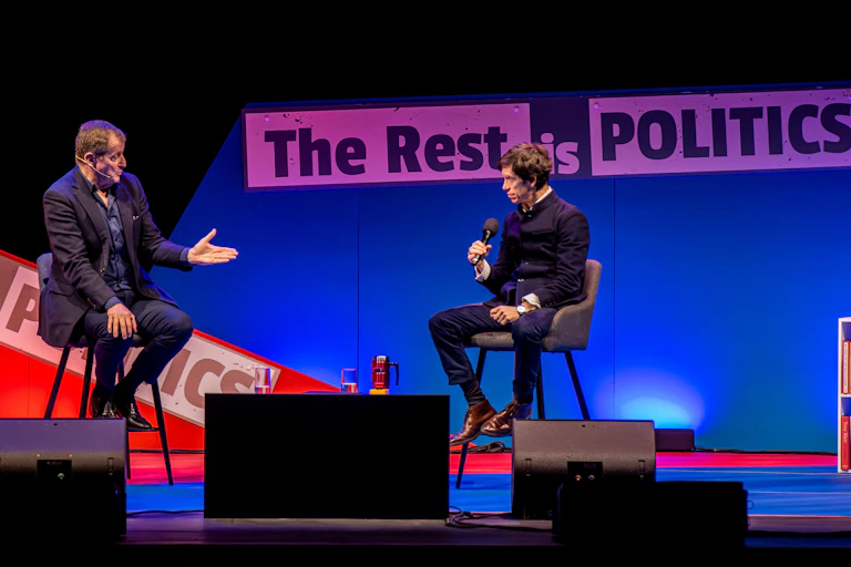 Campbell and Stewart on stage at a live podcast show, with a red and blue background reading The Rest is Politics behind them