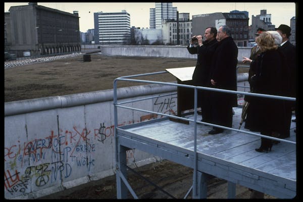 A group of men wearing black jackets stand at the edge of a platform and look toward gray buildings over a wall.