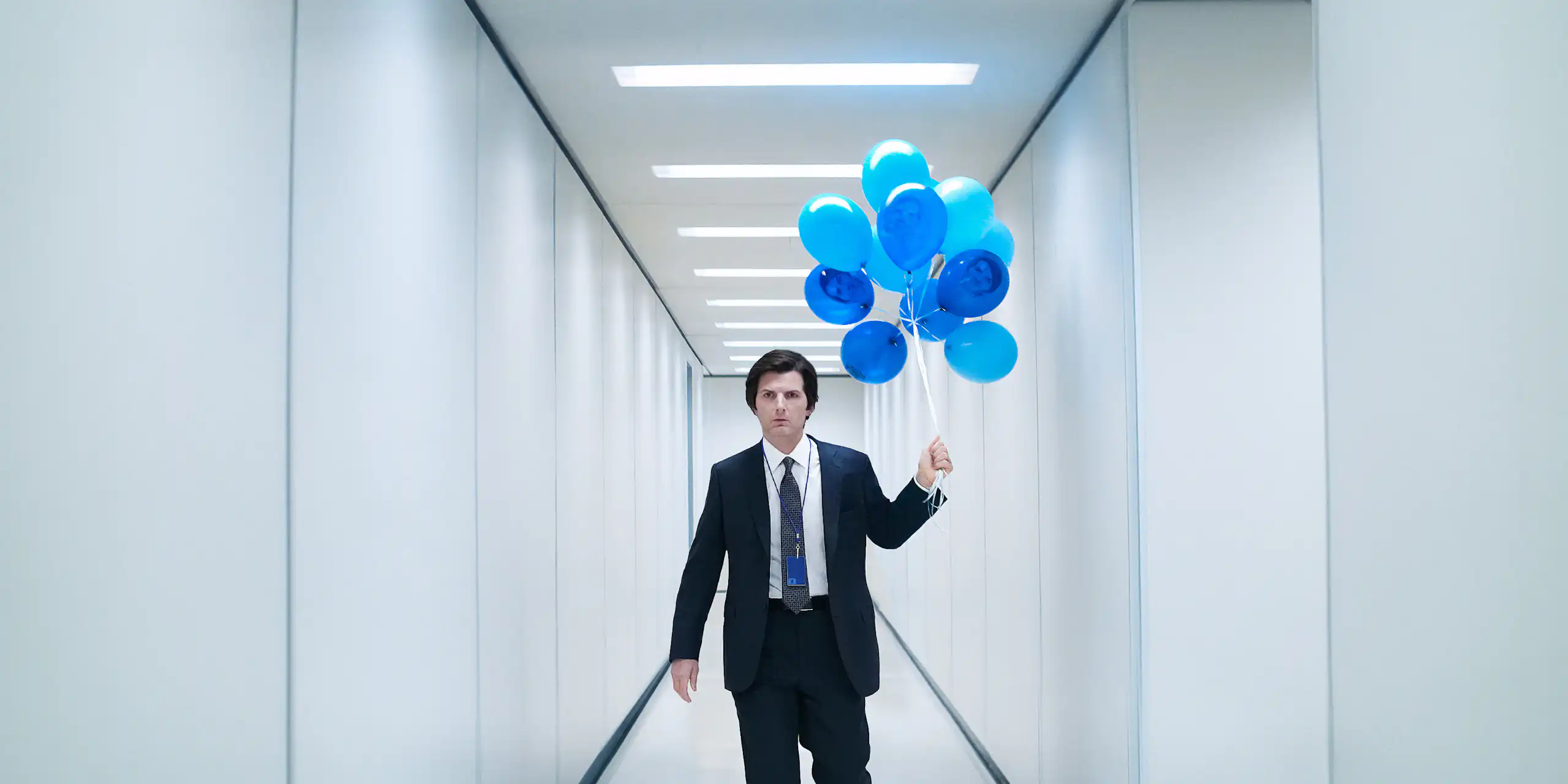 Adam Scott in a white hallway holding blue balloons