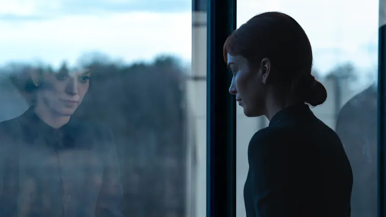 Woman looking out of a window with a sad expression, her reflection mirrored back to her