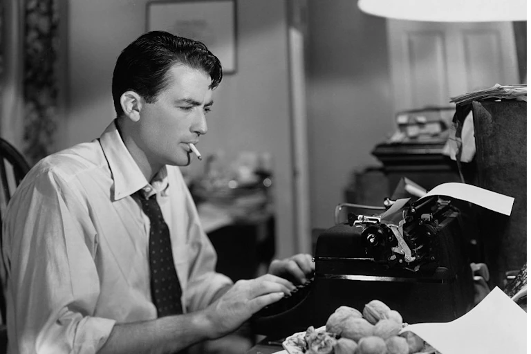 A black and white photo of a man in a shirt and tie using a typewriter while smoking a cigarette.