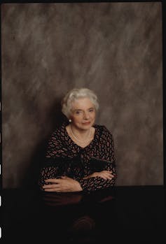 A formally dressed woman with white hair poses, with her arms folded, in front of a marbled backdrop.