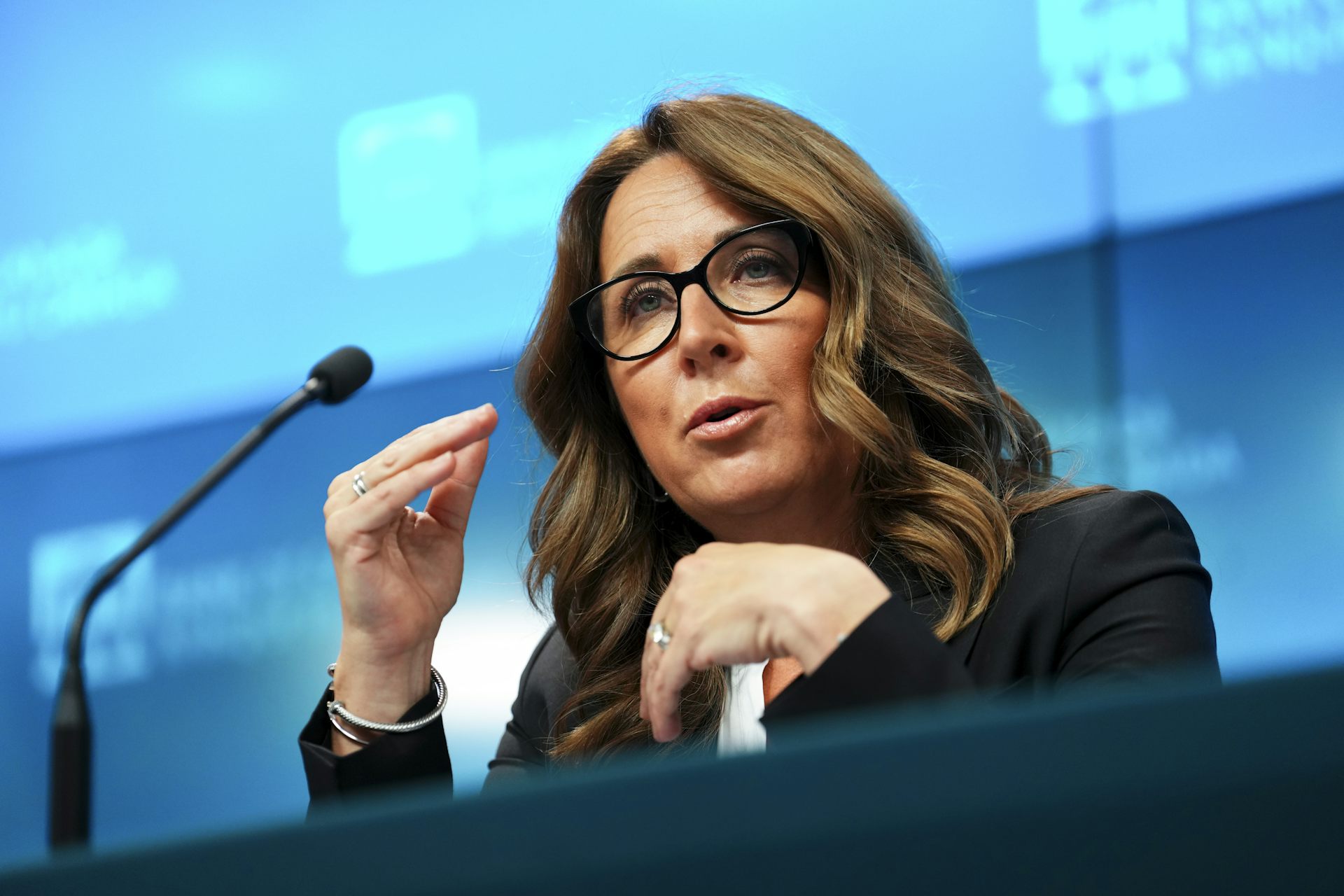 A woman with thick dark hair and glasses speaks into a microphone.