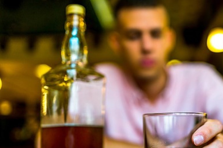 Sitting at a bar with a bottle in front of him, a man holds a drink.