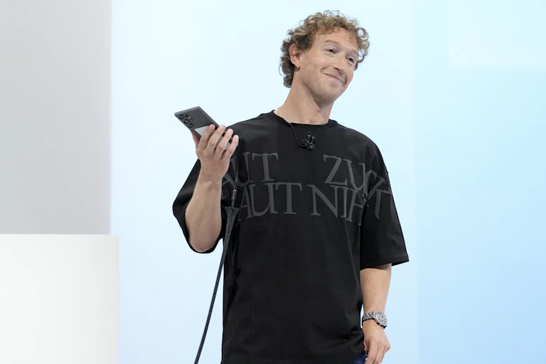 Man with short curly hair standing on stage, holding a phone and smiling.