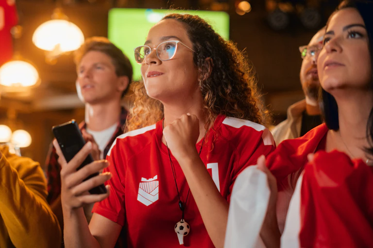 A young woman in a sports jersey holds a smart phone and watches a sports game