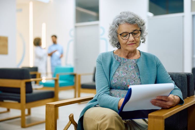 A senior woman in a medical waiting room looking at a clipboard.