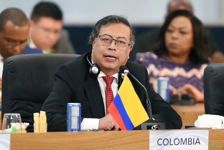 Gustavo Petro hablando sentado ante una mesa con micrófono y la bandera de Colombia.
