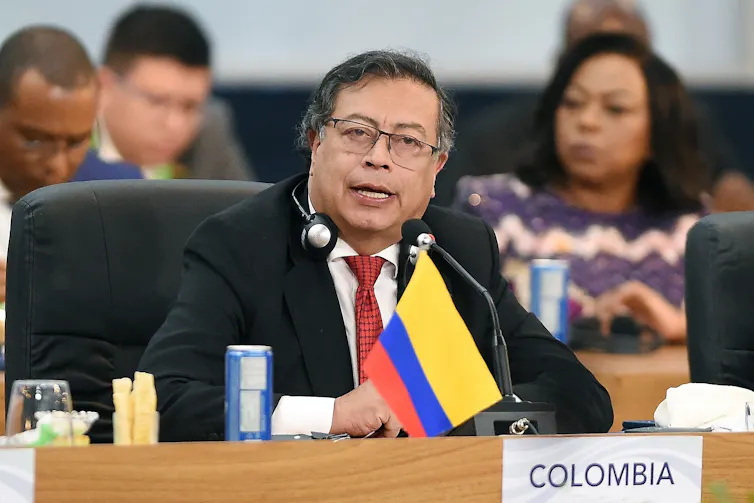Gustavo Petro hablando sentado ante una mesa con micrófono y la bandera de Colombia.