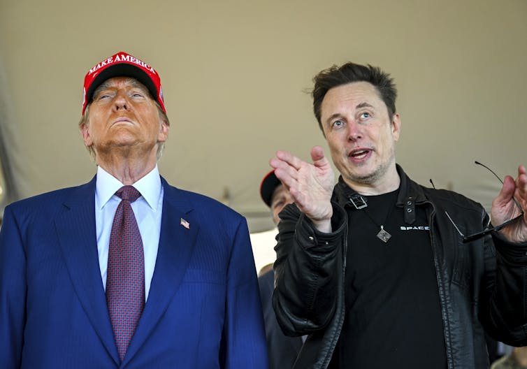 An older man with heavy facial bronzer wearing a red ball cap stands next to a younger man with dark hair who gestures as he looks ahead.