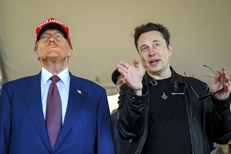 An older man with heavy facial bronzer wearing a red ball cap stands next to a younger man with dark hair who gestures as he looks ahead.
