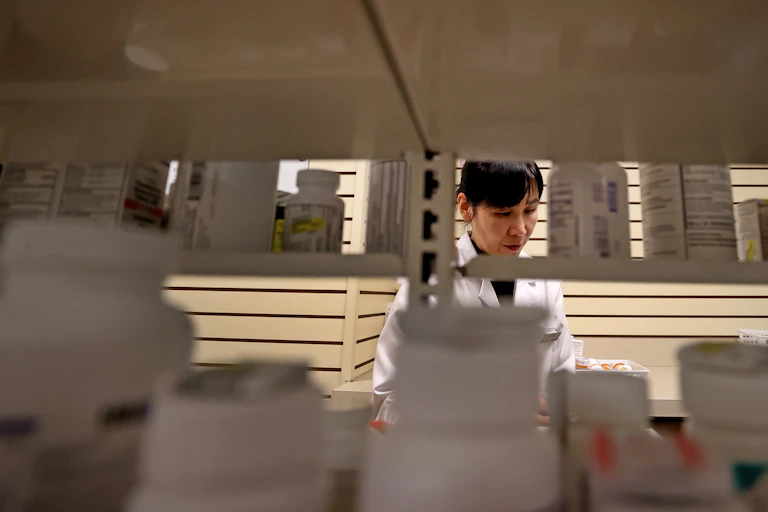 A woman in a white coat looks at a shelf. She is framed by medication bottles.