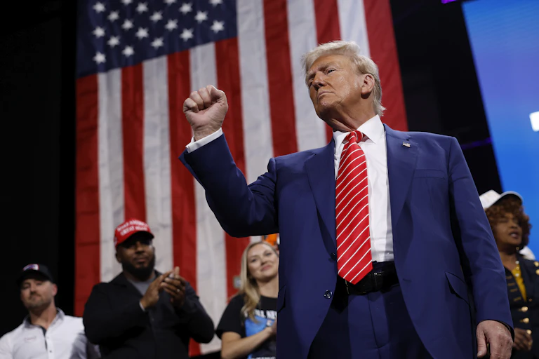 A man in a suit and tie stands with a clenched fist in front of an American flag.