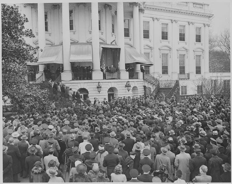 A man stands on the balcony of a large white building speaking to a crowd.