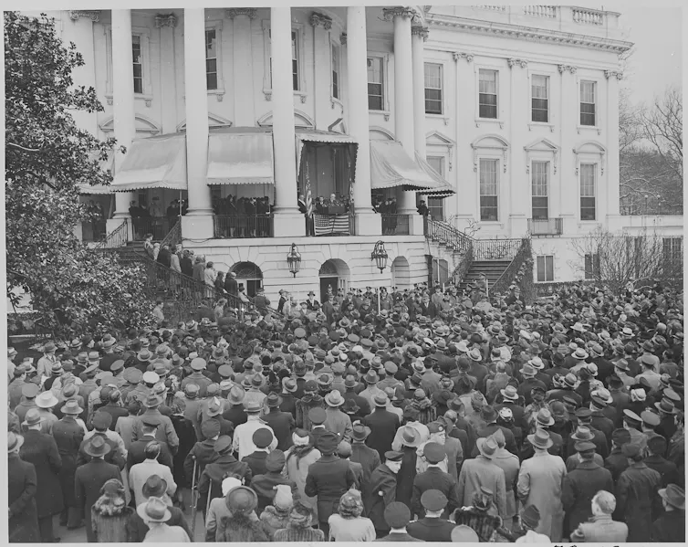 A man stands on the balcony of a large white building speaking to a crowd.