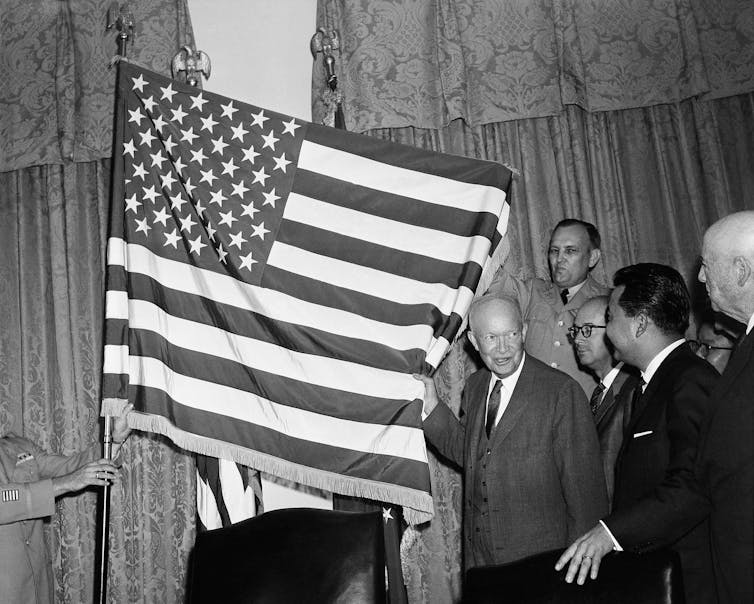 A black-and-white photo shows a man unfurling the U.S. flag with 50 stars.