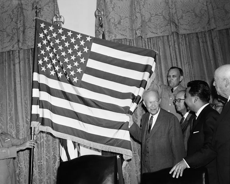 A black-and-white photo shows a man unfurling the U.S. flag with 50 stars.