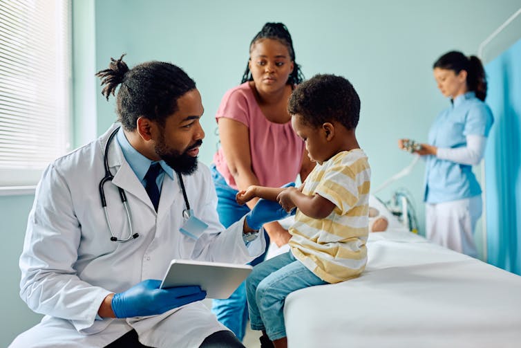 A medical office, with a man in a while coat examining a boy while his mother looks on, and another health-care professional in the background
