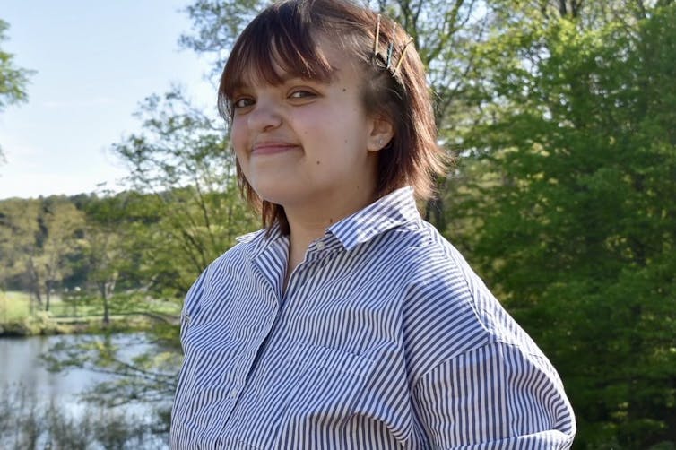 A smiling woman in a triped shirt, standing near a stream.
