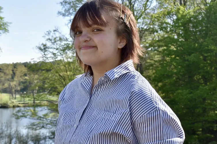 A smiling woman in a triped shirt, standing near a stream.