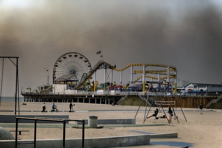 People sit on swings at the beach watching thick dark smoke over the city.