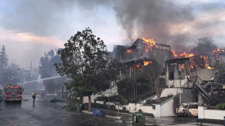 Un camion de pompiers pulvérise de l’eau depuis la rue alors que des maisons brûlent à flanc de colline