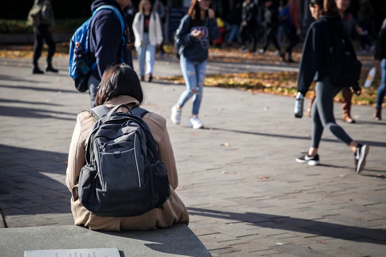 A woman with a backpack sits by a path as other people walk by