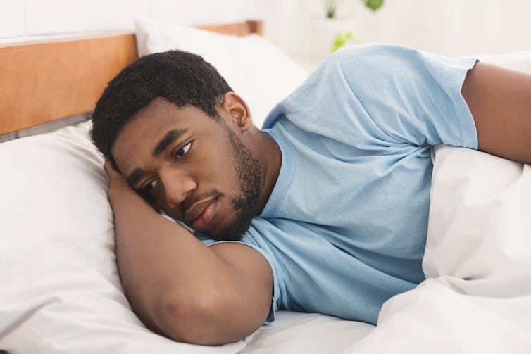 A man wearing a blue t-shirt lying alone in a bed with a pensive expression