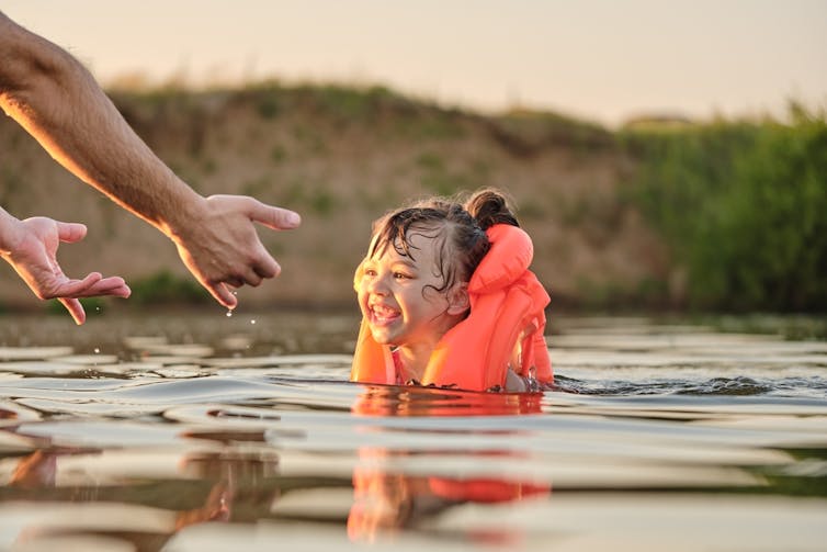 A happy small girl in a bright orange vest laughs while immersed in a river.