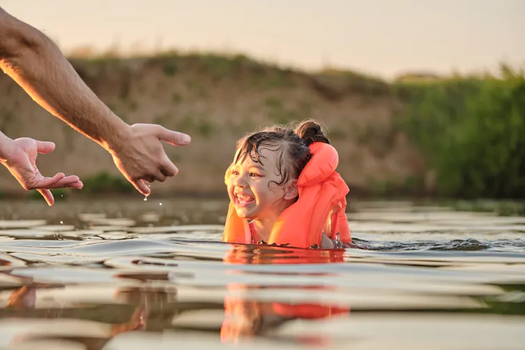 A happy small girl in a bright orange vest laughs while immersed in a river.