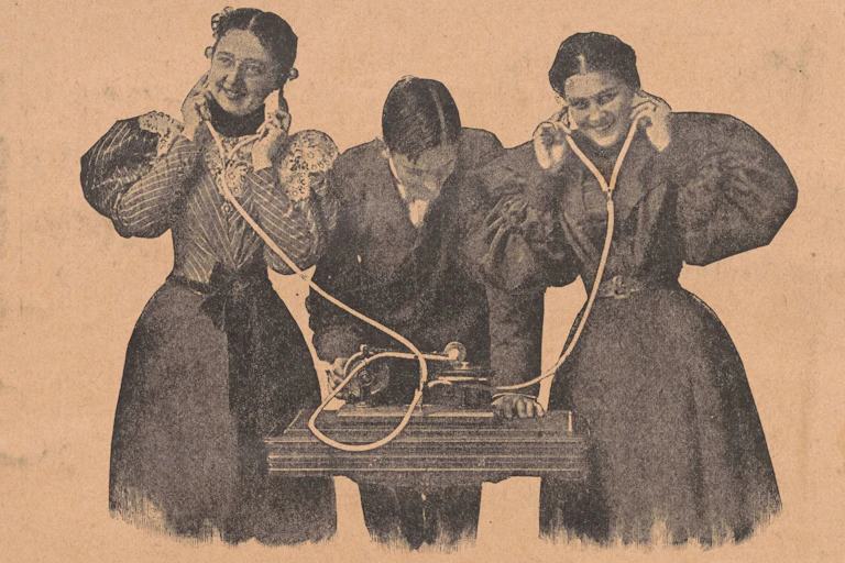Two women and a man. The women listen to a gramaphone with headphones.