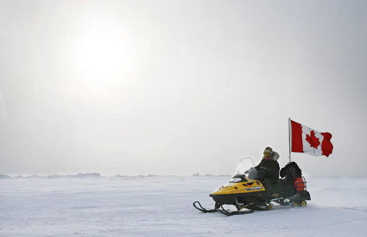 A snowmobile with a Canadian flag on the back drives across snow