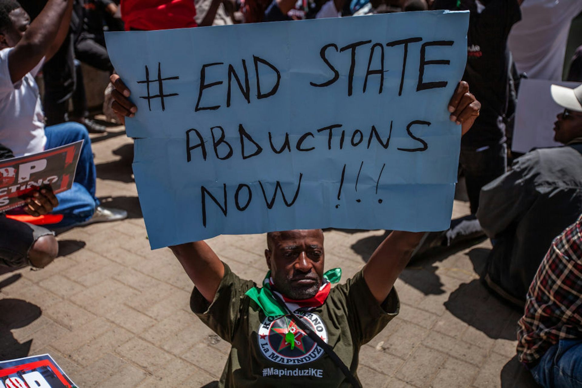 A man seating the ground holding up a manila paper with the words '#End state abductions now!!!'