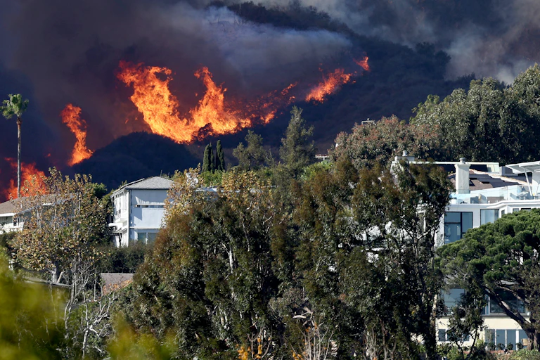 Fires burn in the hills behind Pacific Palisades houses with dry vegetation surrounding them.