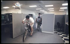 Dos hombres montan bicicletas a través de un laberinto de cubículos. Un globo plateado con forma de estrella se eleva de un cubículo.